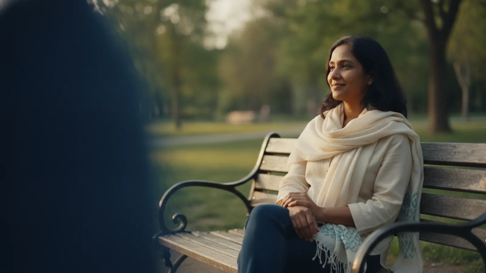 A woman sits at ease on a park bench in warm afternoon light, hands resting in her lap.