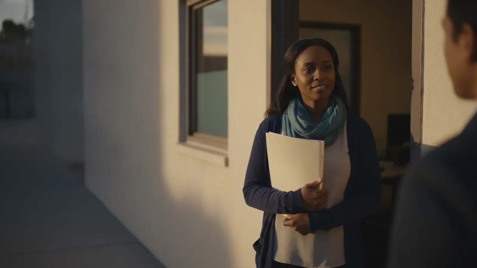 A broker stands in an office doorway in warm afternoon light, a closed folder held against her, mid-conversation.
