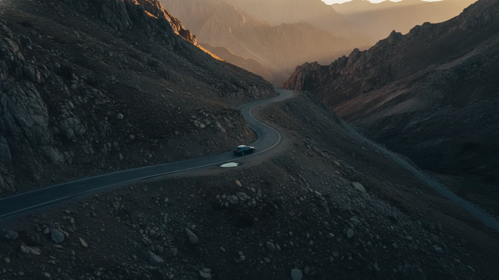 A single car on a mountain road at dusk, seen from a viewpoint above with warm light along the ridges.