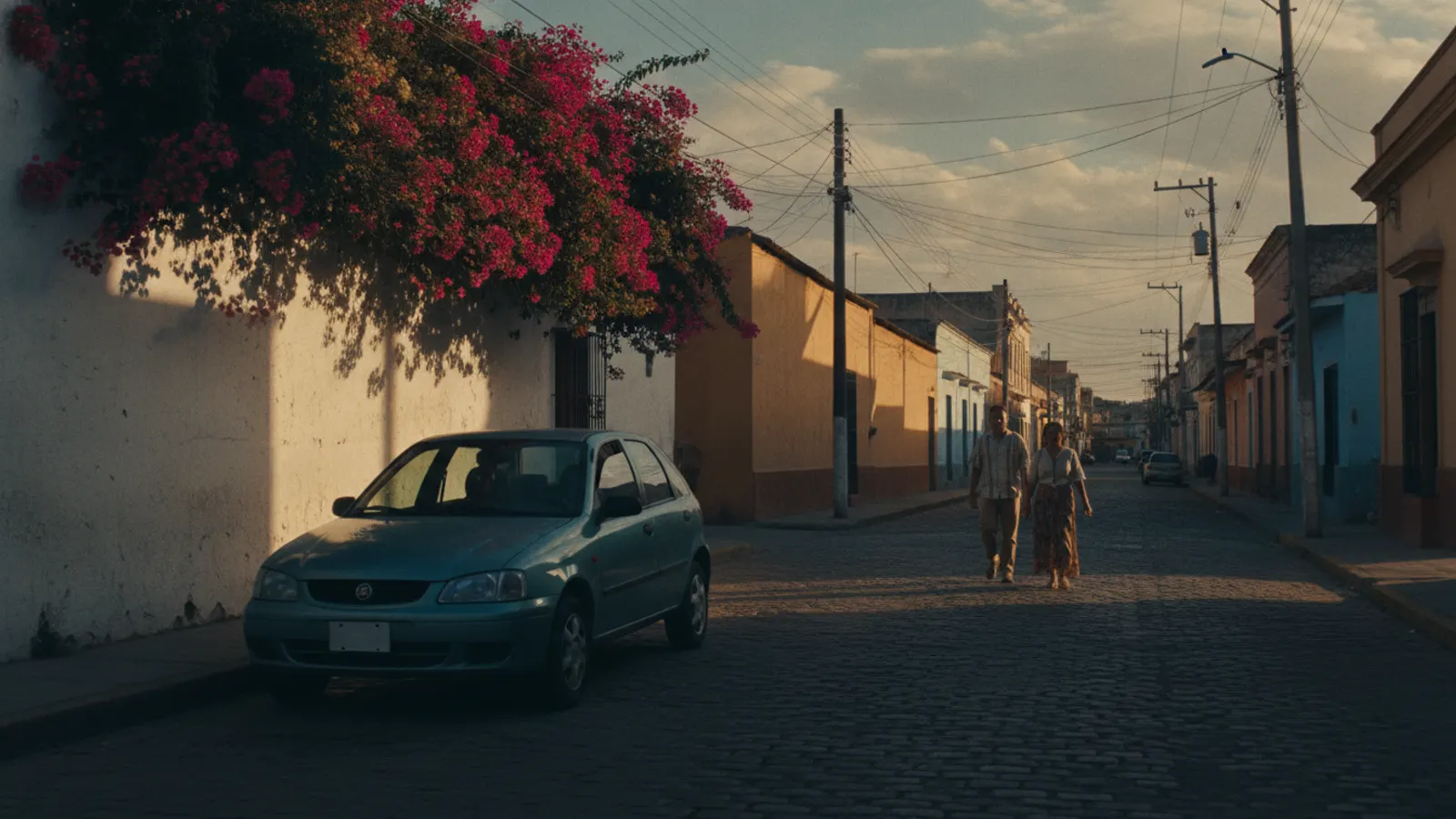 A sunlit Latin American street in late afternoon, with painted concrete walls and a car parked in the shade.